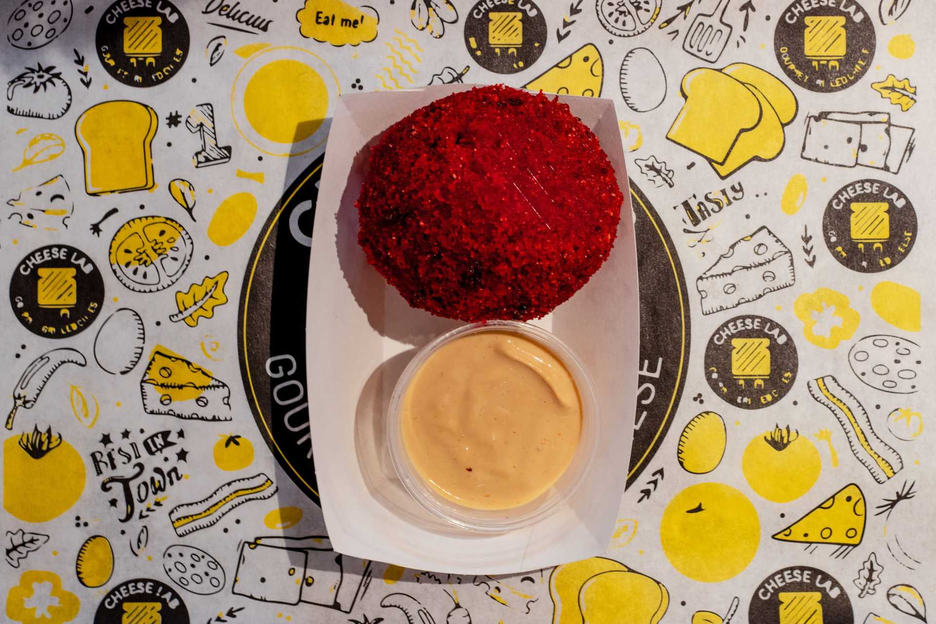 Red breaded cheese ball with dipping sauce on cheese-themed paper.